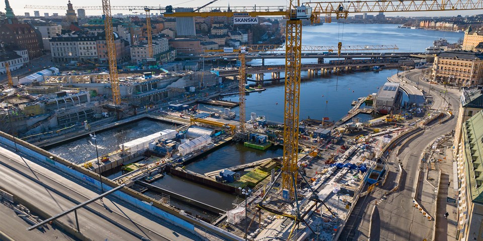 Drönarfoto över arbetsområdet vid Slussen. Vy från nordväst över vattenområdet där fyra gula tornkranar står och byggarbeten pågår. 